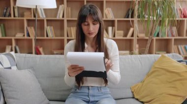 Elinde belge tutan, odaklanmış bakışlarıyla ileriye bakan, ciddi derecede çekici, beyaz bir kadın. Kitap rafları ve yumuşak ışıkla sakin bir odada otururken raporun analiz edilmesi veya tartışmaya hazırlanması