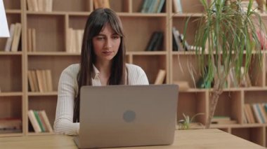 Ciddi derecede güzel beyaz bir kadın dizüstü bilgisayarı kapatıyor ve aydınlık odada bitkin görünüyor. Uzun bir çalışma seansından sonra ara vermek ya da ahşap kitaplığın yanında otururken çevrimiçi toplantıyı bitirmek.