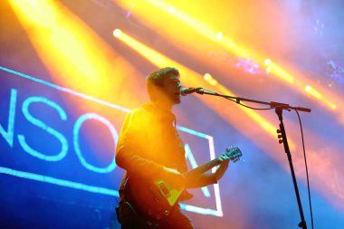 Sahnede, renkli spot ışıkları ve mikrofon altındaki bir konserde elektro gitar çalıp şarkı söyleyen rock gitaristi.