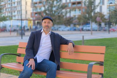 Parktaki bankta oturan adam sakin ve kasketli gülümsüyor.