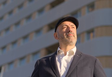 Şık şapkalı bir adamın portresi gururla şehrin arka planında binalarla kente bakıyor.