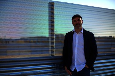 Zarif, olgun bir adam sakin bir şekilde endüstriyel şehir ortamında kameraya bakıyor.