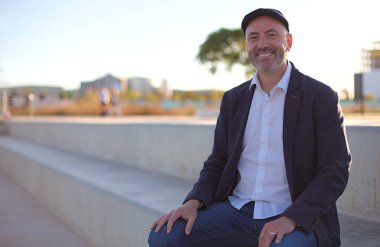 Neşeli orta yaşlı bir adam günlük kıyafetlerle beton bir bankta oturuyor açık bir şehir ortamında, güneşli bir günde arka planda bulanık bir şehir manzarası.