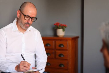 A therapist listens attentively and takes notes during a confidential session with a mature female client in a modern, private office clipart