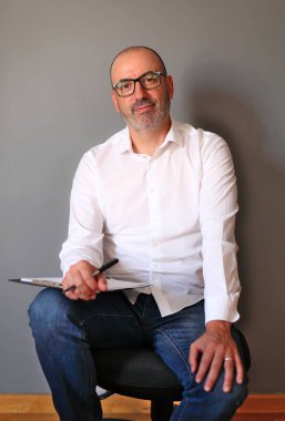 Rahat bir pozisyonda, belgelerini incelerken iş kıyafetleri giyen olgun bir birey. Görüntü profesyonelliği, güveni ve gelişigüzel ama odaklanmış bir ambiyansı yansıtıyor..