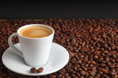 Koyu arkaplanda kavrulmuş kahve çekirdekleri ile çevrili espresso dolu beyaz bir kahve fincanı.