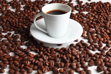 Temiz bir yüzeyde kavrulmuş fasulyelerle çevrili sıcak siyah espresso kahvesiyle dolu beyaz bir fincan sıcaklığı ve zengin aromayı ön plana çıkarıyor.