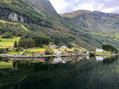 A village surrounded by cliffs reflecting in calm waters along Naeroyfjord, Norway's narrowest fjord. Traditional wooden houses with red, white, and brown hues and small white church are scattered on lush, green terrain.  clipart