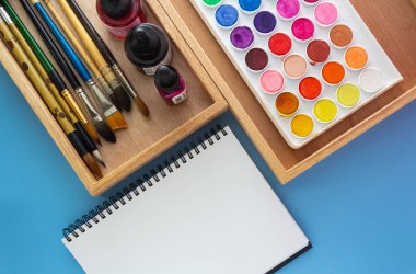 Mavi arkaplanda beyaz çizim defteriyle suluboya boya araç kutusu.