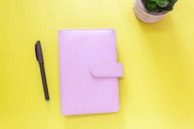 Manyetik tokası, siyah kalemi ve canlı sarı arka planda sulu bitki saksı köşesi olan pastel pembe deri bir planlamacı içeren minimalist düz bir kompozisyon. Planlama ve organizasyon kavramları için temiz modern kurulum ideali.