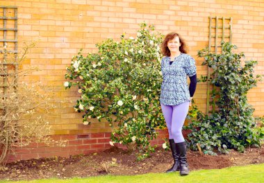 A glamourous brunette lady is pictured proudly standing beside camelias just coming into flower in early March. clipart