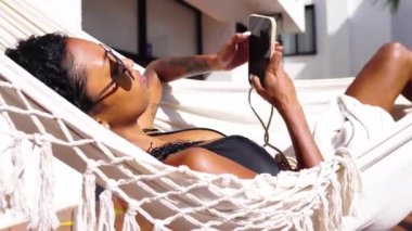 Bikini 'deki genç siyahi kadın sıcak güneşli bir günde hamakta ellerinde akıllı telefonuyla yatıyor.