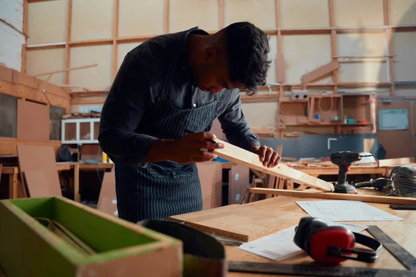 Multiracial young man examining wood plank while holding pencil at woodworking factory