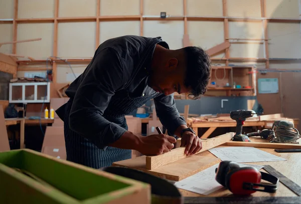 木工工場の電動工具の横に木板に鉛筆で書く多人種の若い男 — ストック写真