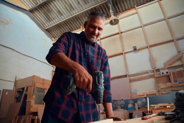 木工工場の木杭の横の板にドリルを使用しながら笑顔の多人種中大人の男 — ストック写真