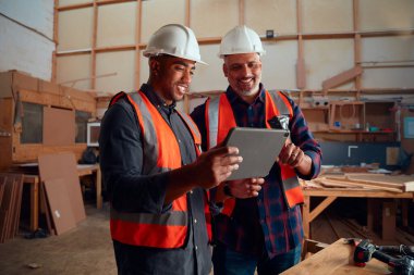 Yansıtıcı yelek ve kasklı çok ırklı adamlar marangozluk fabrikasında gülümserken dijital tablet tutuyorlar.