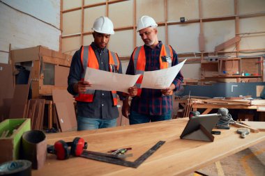 Dijital tablet ve ahşap fabrikasındaki aletlerin yakınındaki kağıtları inceleyen yansıtıcı yelek giyen çok ırklı adamlar.