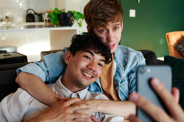 年轻的同性恋夫妇在家里手牵手坐在沙发上 一边用手机自拍 — 图库照片