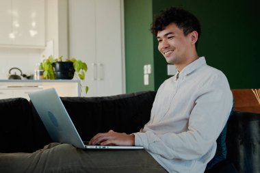 Evdeki oturma odasında ayakları kanepede otururken dizüstü bilgisayar kullanan tişörtlü mutlu genç adam.