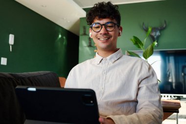 Evdeki kanepede kablosuz kulaklıkla dijital tablet kullanırken gülümseyen tişörtlü çok kültürlü genç adam.