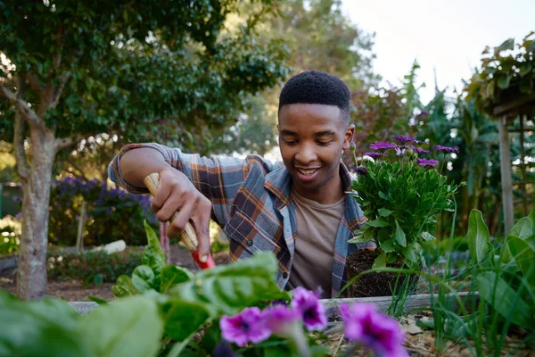 Happy young black man in checked shirt using spade in flowerbed while gardening in garden center