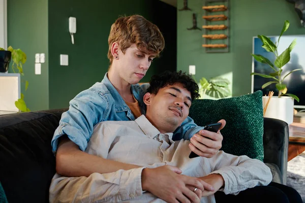 Young Gay Couple Using Mobile Phone While Embracing Sofa Living — Stock Photo, Image