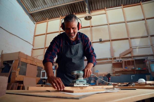 耳のマフを身に着けている間に笑顔の多人種中大人の男と木工工場で木材の上の電動工具を使用して — ストック写真