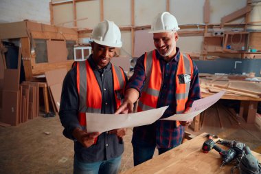 Yansıtıcı yelek giymiş çok ırklı adamlar marangozluk fabrikasındaki elektrikli aletlerin yanındaki kağıda gülümsüyor ve işaret ediyorlar.