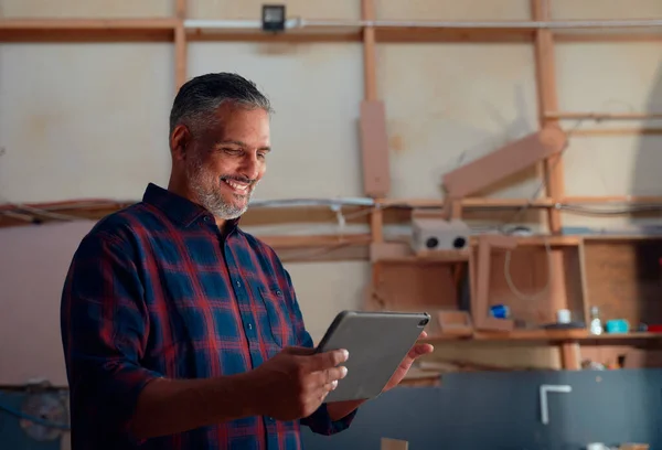 Multiracial mid adult man in checked shirt smiling while typing on digital tablet in woodworking factory