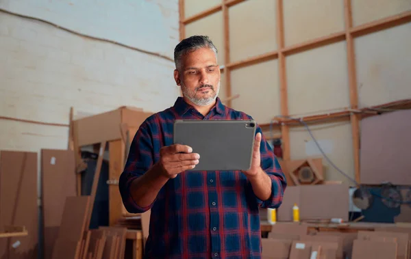 Focused multiracial mid adult man in checked shirt using digital tablet at woodworking factory