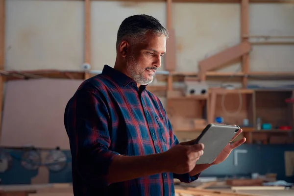Multiracial mid adult man in checked shirt smiling while using digital tablet at woodworking factory