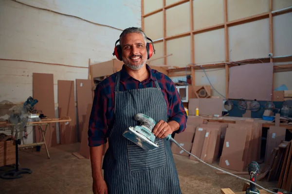 耳のマフを身に着けている間に笑顔の多人種中大人の男と木工工場で電動工具を保持 — ストック写真