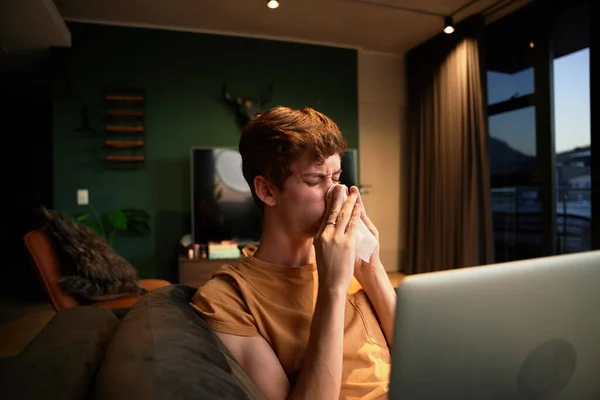 Ill young caucasian man in casual clothing blowing nose next to laptop on sofa in living room at home