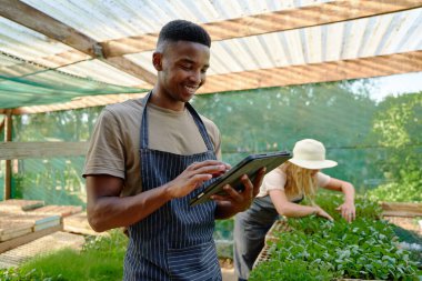 Young multiracial couple in aprons smiling and using digital tablet while gardening in plant nursery