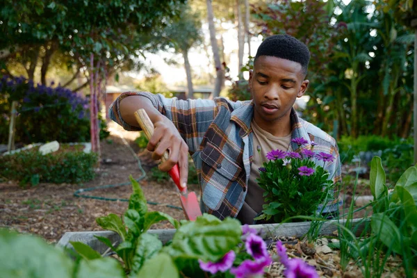 Young black man in checked shirt using spade in flowerbed while gardening in garden center