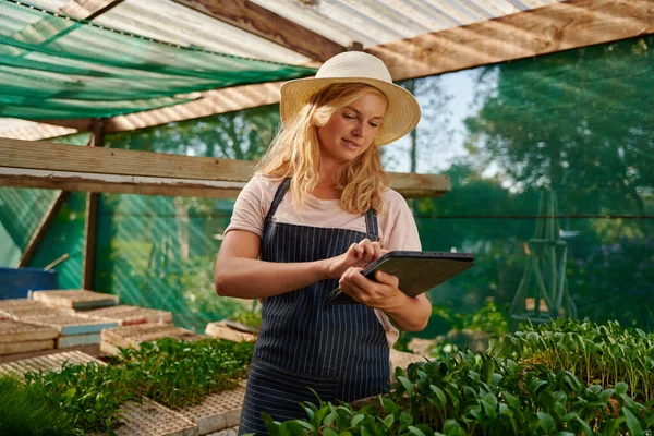 Young caucasian woman in apron smiling while using digital tablet next to plants in plant nursery