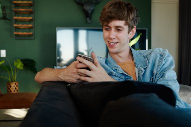 Happy young caucasian man in casual clothing smiling while texting on mobile phone in living room at home