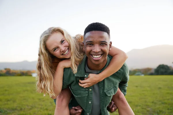 Happy multiracial young couple smiling and looking at camera during playful piggyback in field