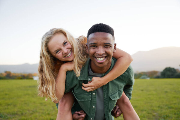 Happy multiracial young couple smiling and looking at camera during playful piggyback in field