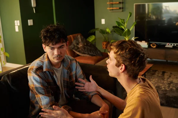 Frustrated young gay couple wearing casual clothing arguing and gesturing in living room at home
