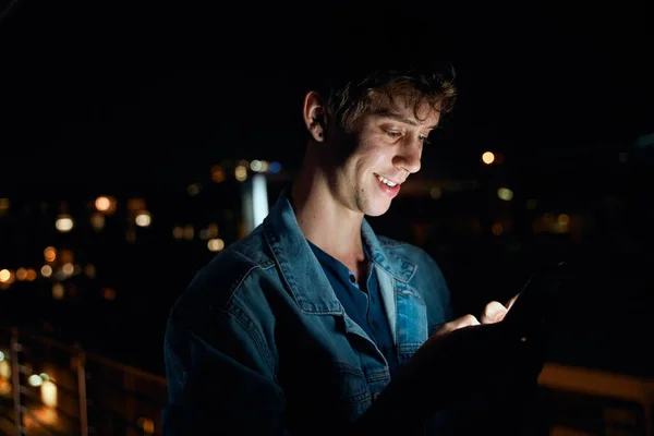 Young caucasian man in casual clothing smiling while typing on mobile phone on balcony at night