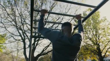 Rear view of young black man doing chin-ups and stretching by the monkey bars in the park