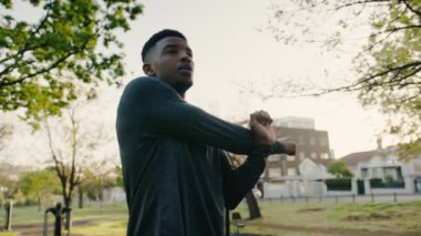 Spor elbiseli atletik genç siyah adam parkta ağaçların arasında esneme hareketleri yaparken gülümsüyor.