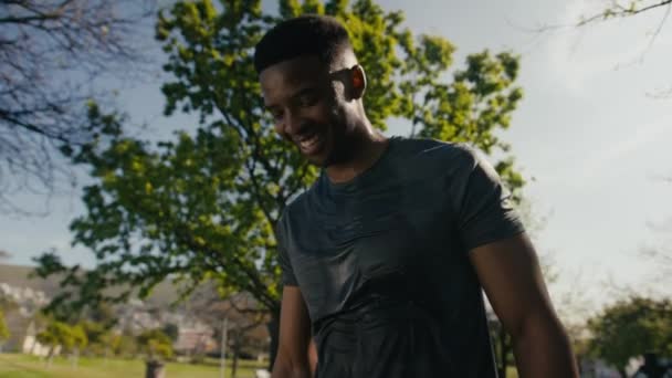 公園で木のそばで休憩や水を飲みながら笑顔でTシャツを着たアスレチックな若い黒人男性 — ストック動画