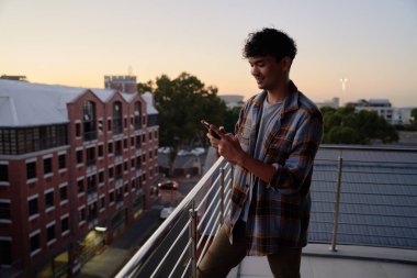 Tişörtlü genç adam gün batımında apartmanın balkonunda telefon kullanıyor.