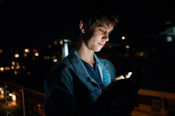 Young caucasian man in casual clothing texting on mobile phone outside apartment at night