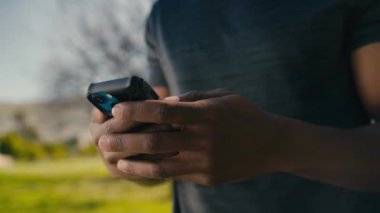 Genç siyahi adamın cep telefonuyla yazışması ve parkta mola verirken gülümsemesi.