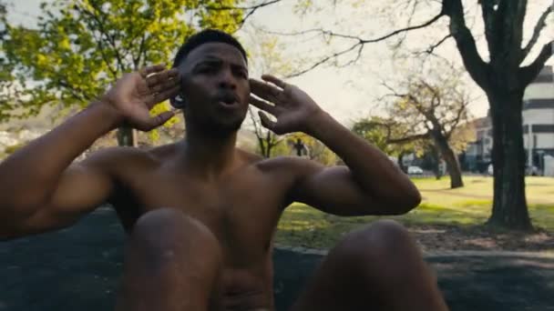 無線ヘッドフォンを持つトップレス若い黒人男性が公園の歩道で座り込みをしています — ストック動画