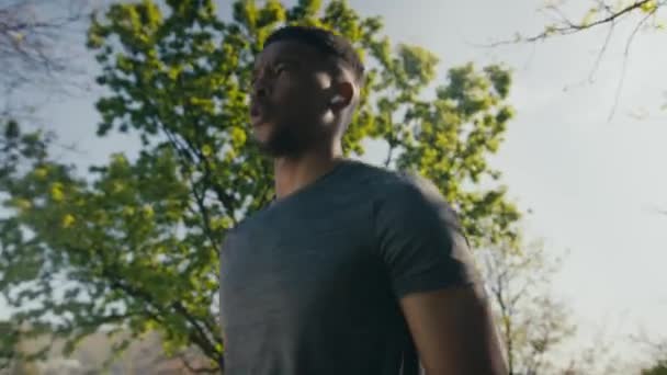 公園でフィットネストレーニング中に肺を行うTシャツとワイヤレスヘッドフォンのアスレチック若い黒人男性 — ストック動画