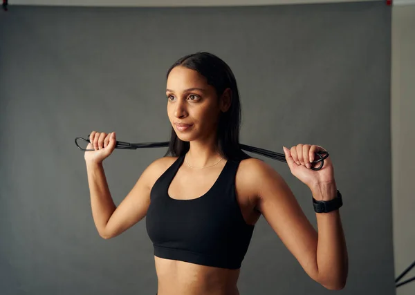 Determined young biracial woman wearing sports bra holding jump rope behind neck in studio